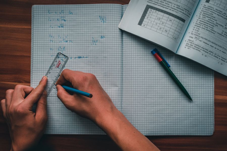 Deux mains tracent une figure à la règle sur du papier quadrillé, à côté de formules écrites au stylo bleu.
